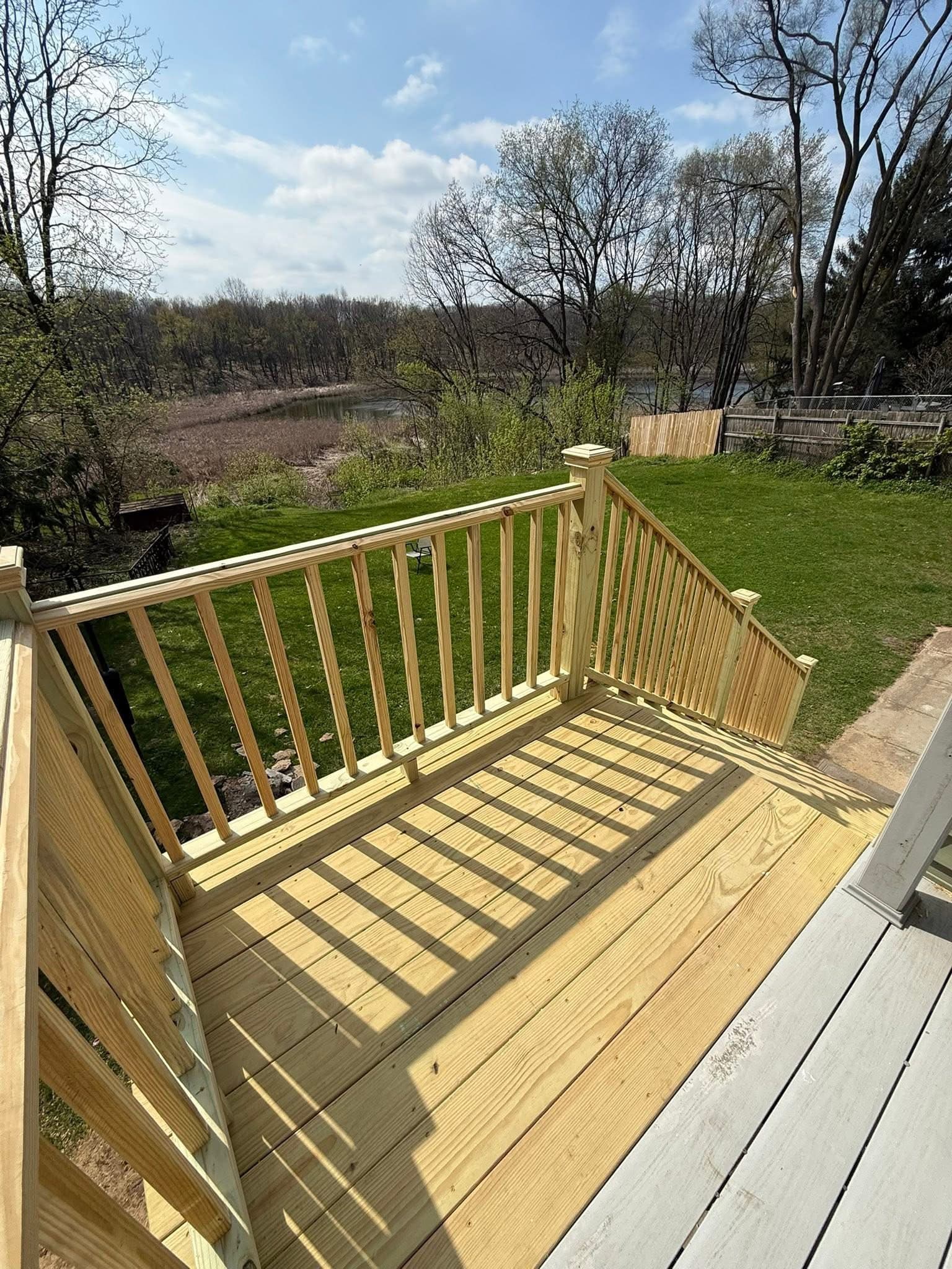 Wooden deck with railings overlooking a green lawn and a distant lake under sunny skies.