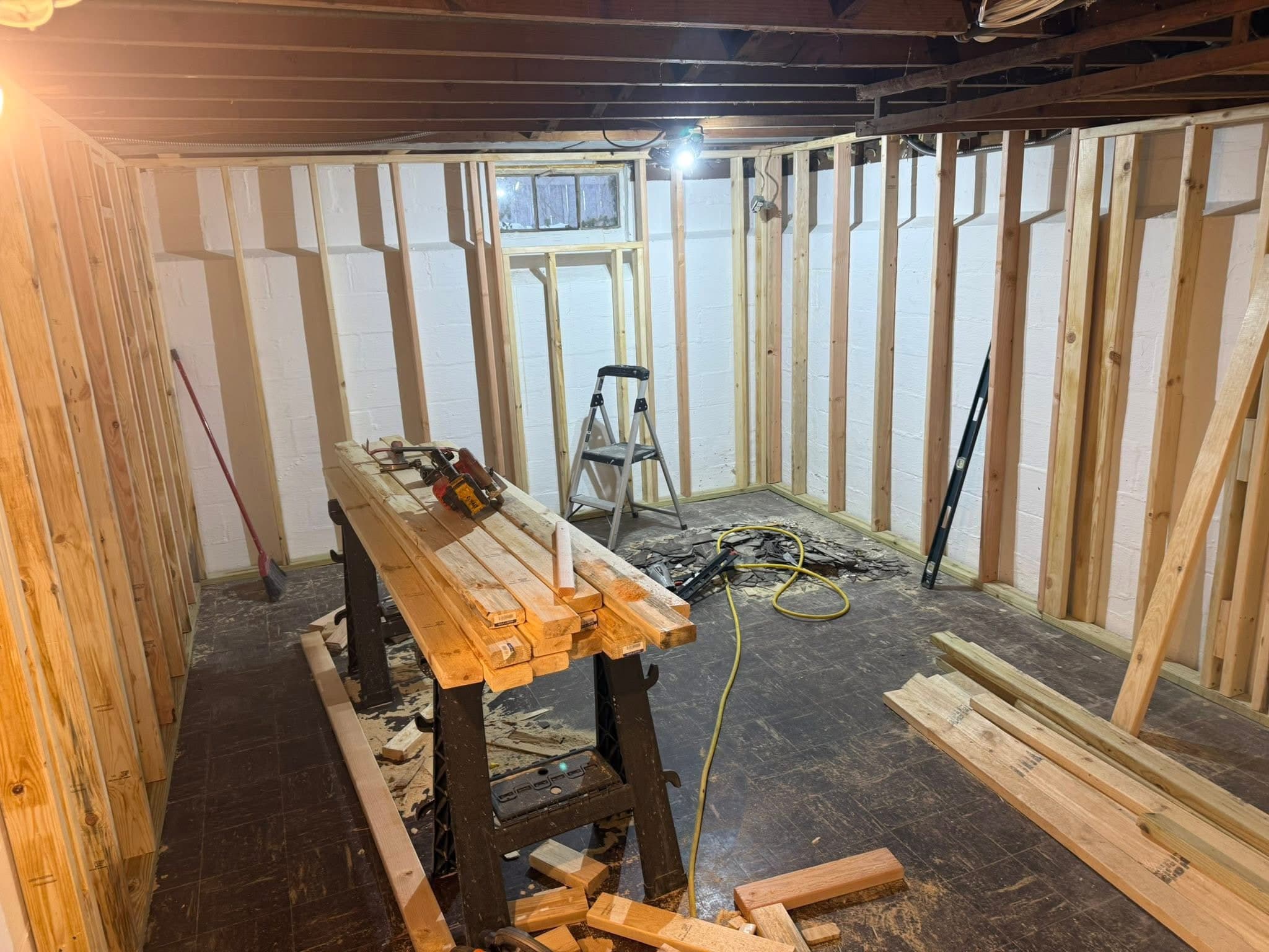 Unfinished basement with wooden wall studs, a sawhorse workbench, and various construction tools.