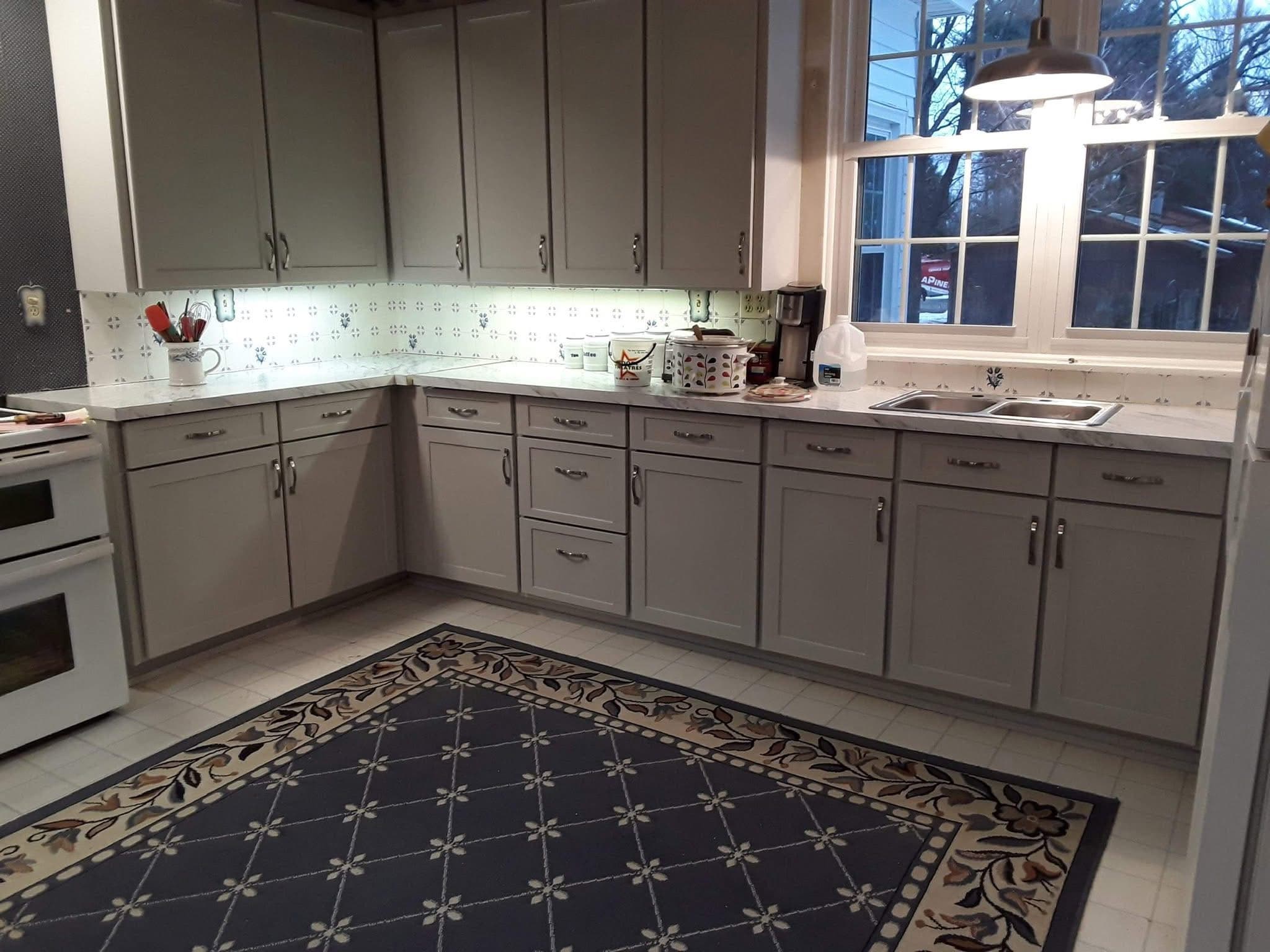 Light gray kitchen cabinets, white countertops, under-cabinet lighting, and a large blue patterned rug.