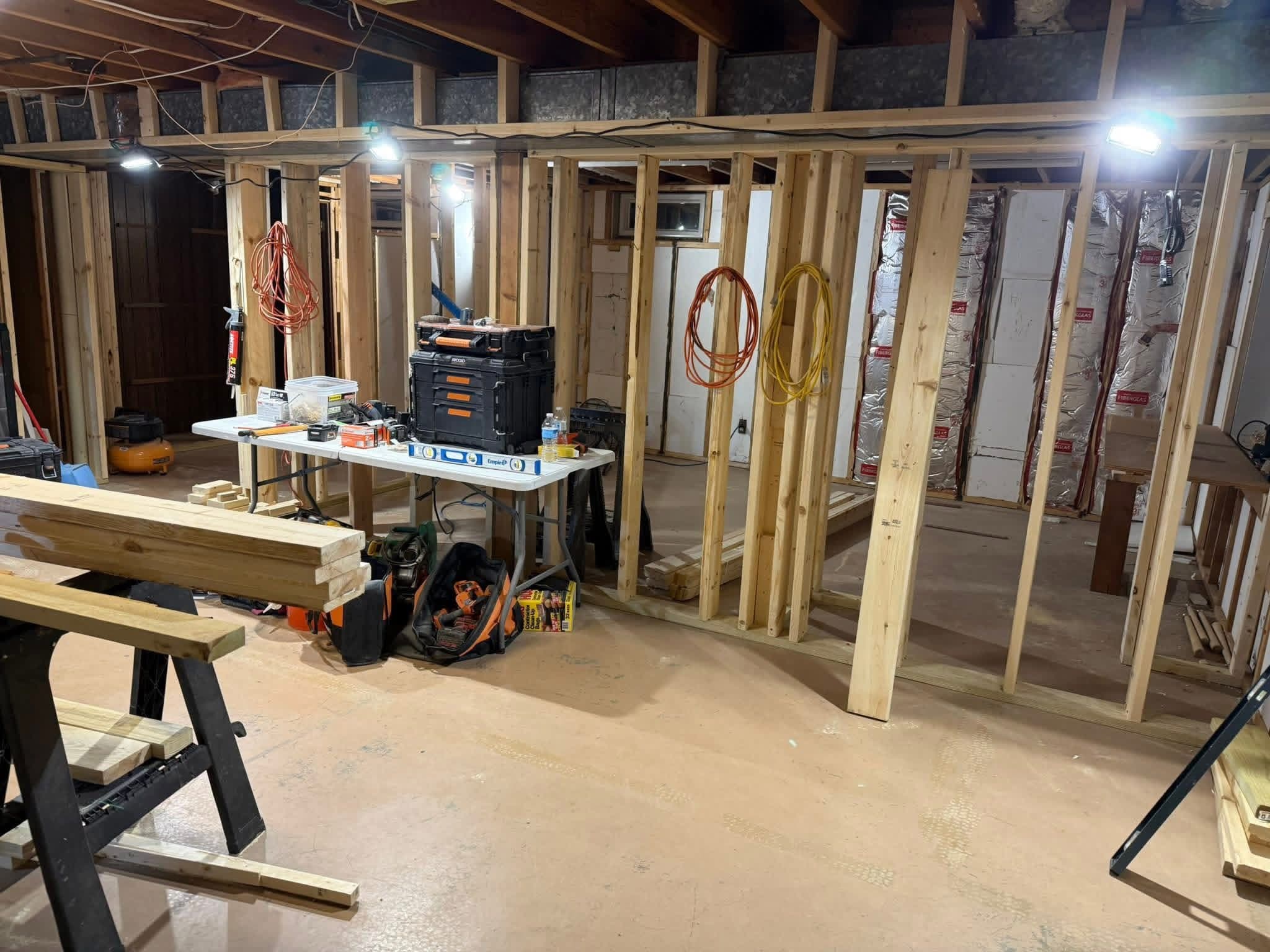 Unfinished basement construction zone featuring wooden wall studs, toolboxes, and bright work lights.