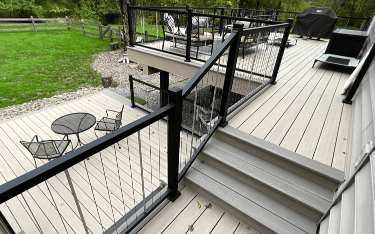 Light gray multi-level deck with black cable railings, stairs, and patio furniture overlooking a lawn.