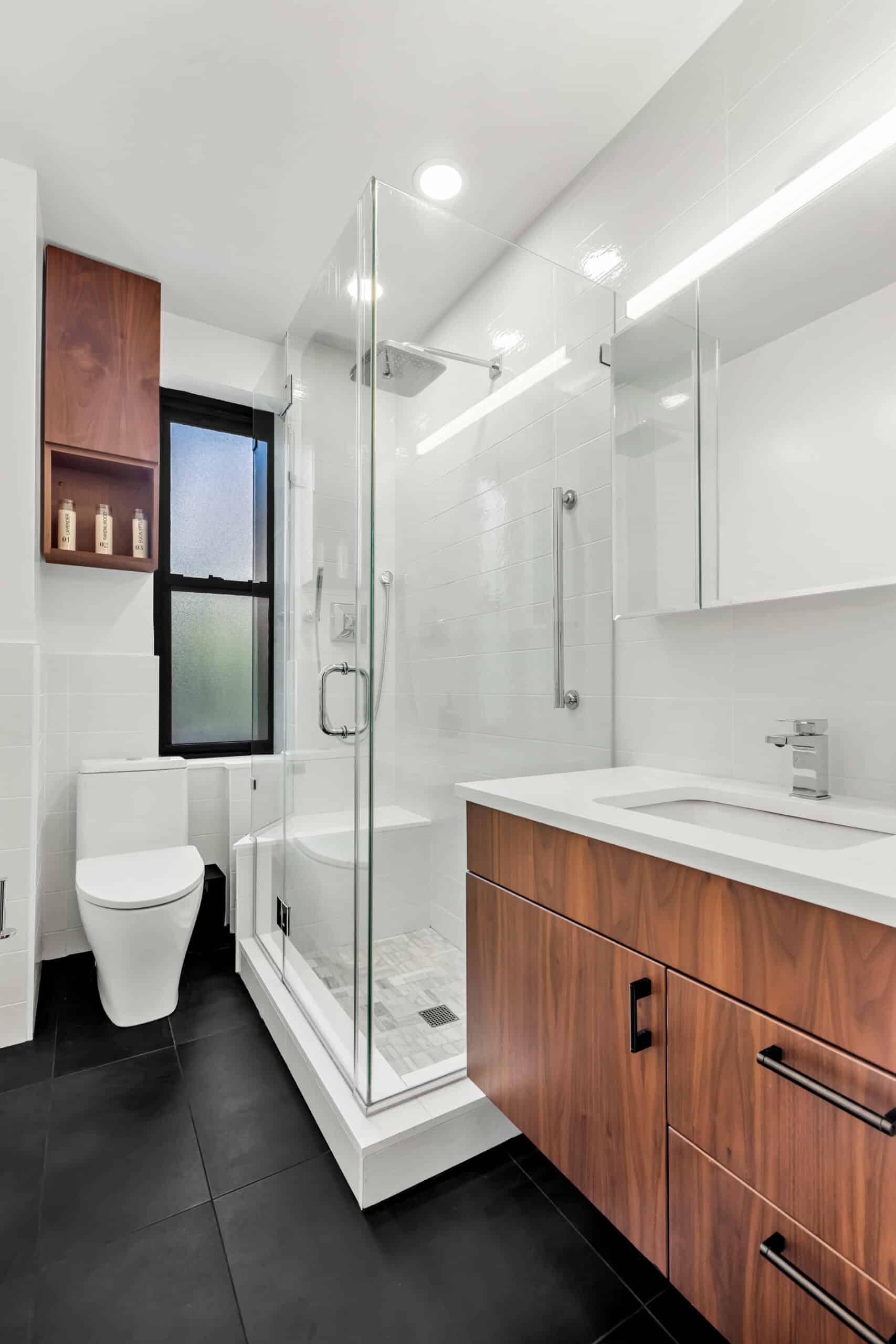 Modern bathroom with glass walk-in shower, wooden vanity, white toilet, and black tile floor.